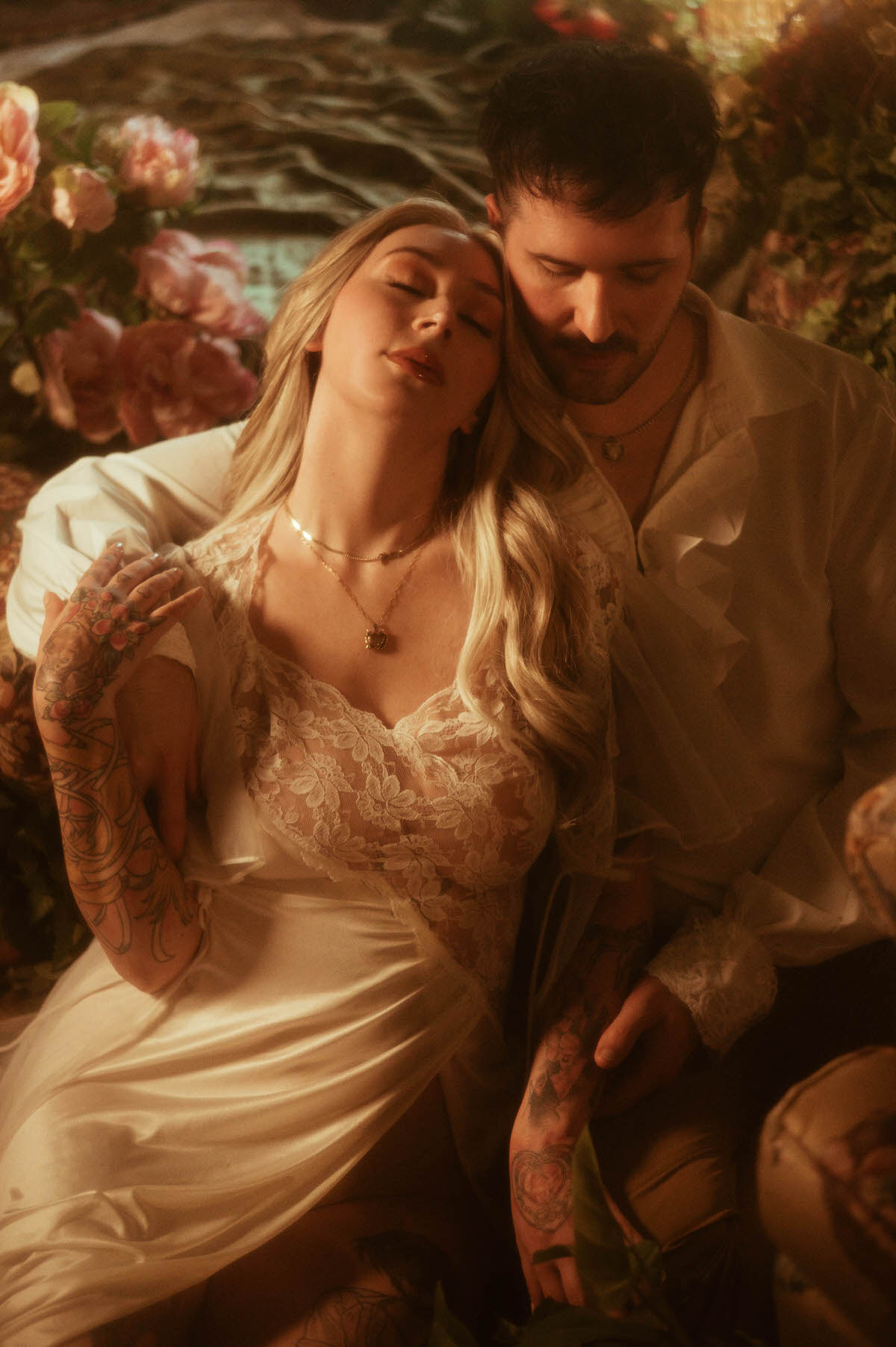 Romantic moment from a Dallas engagement session at Royal Lune Studio, featuring a couple seated among lush florals. The bride-to-be leans back into her partner with eyes closed, wearing a sheer lace slip and gold necklace, while his hand gently cradles her arm, creating a dreamy and intimate spring-inspired portrait.