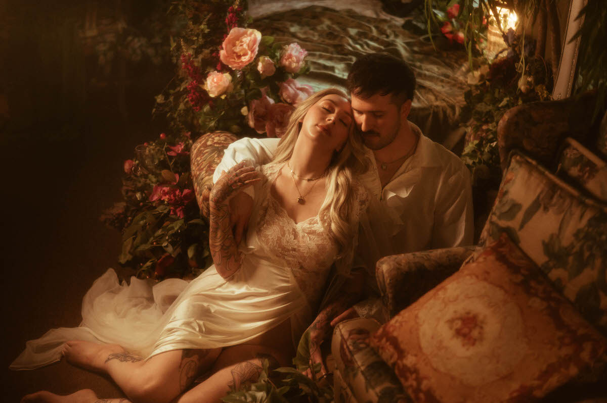 A dreamy engagement portrait taken at Royal Lune Studio in Dallas, featuring a barefoot couple surrounded by lush florals and vintage decor. The woman leans back into her partner’s embrace, wearing a satin and lace slip with her eyes closed, evoking a romantic and painterly atmosphere.