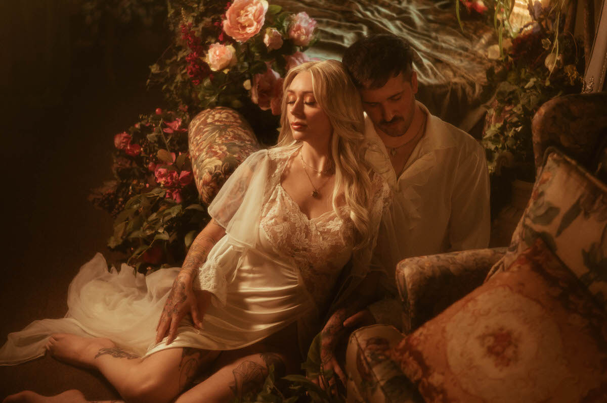 Romantic Dallas engagement photo taken at Royal Lune Studio, showing a barefoot couple nestled among vintage florals and antique furniture. The woman reclines in a lace and satin gown with her eyes closed, as her partner rests his head gently against hers, evoking a tender and intimate fairytale moment.