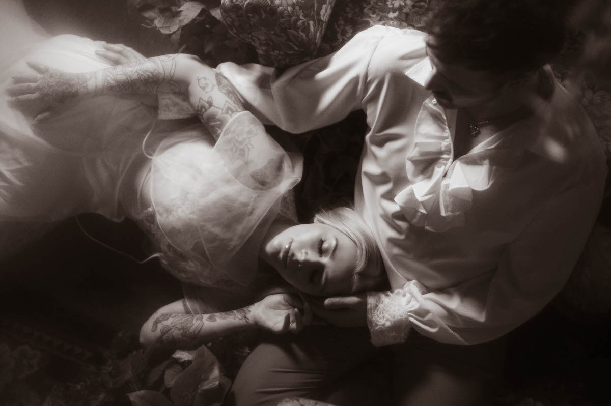 Black and white engagement portrait at Royal Lune Studio, showing the woman lying peacefully with her head in her partner’s lap as he gazes down at her. Her vintage lace gown and floral tattoos add texture and intimacy to the soft, romantic scene.