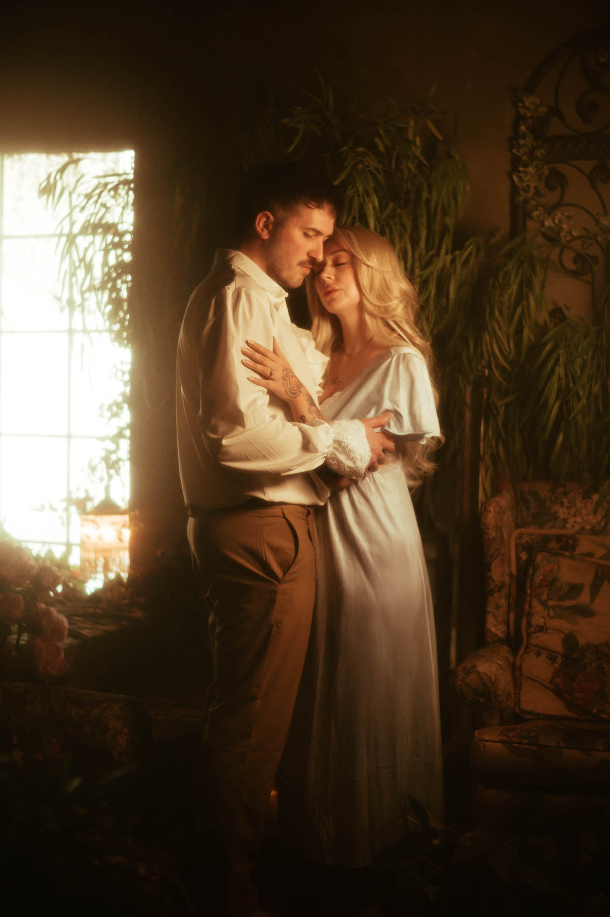 A romantic full-body portrait of a couple embracing in soft window light at Royal Lune Studio, surrounded by vintage furniture, lush greenery, and warm golden tones that evoke a dreamy, timeless engagement moment.