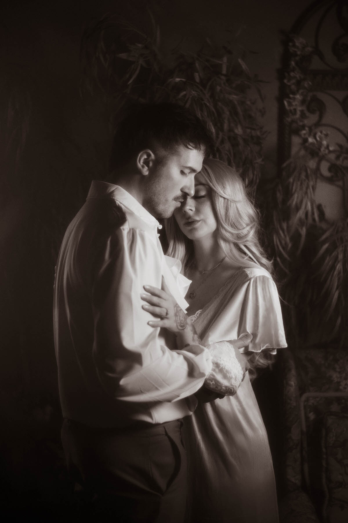 A romantic black-and-white portrait of a couple embracing at Royal Lune Studio, with the woman resting her head tenderly against her partner’s chest, surrounded by vintage decor and soft shadows.