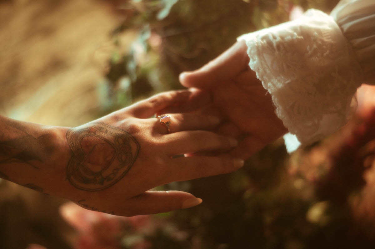 Close-up of the couple's hands at Royal Lune Studio, softly touching as sunlight highlights a gemstone engagement ring and a heart-shaped tattoo, capturing a tender and symbolic moment of their love story.