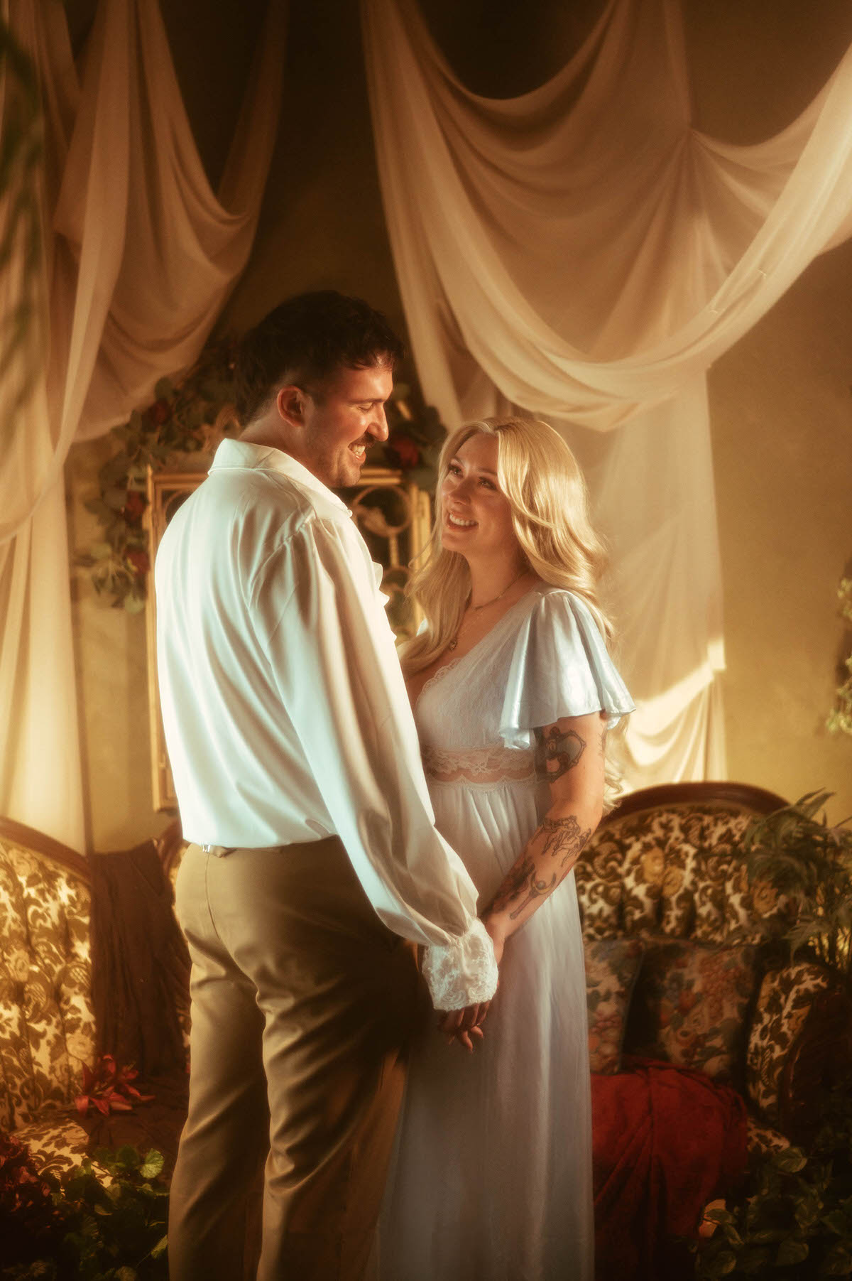 A joyful couple smiles at each other while holding hands in a romantic Dallas engagement session at Royal Lune Studio, surrounded by antique furniture, draped ivory fabric, and golden afternoon light.
