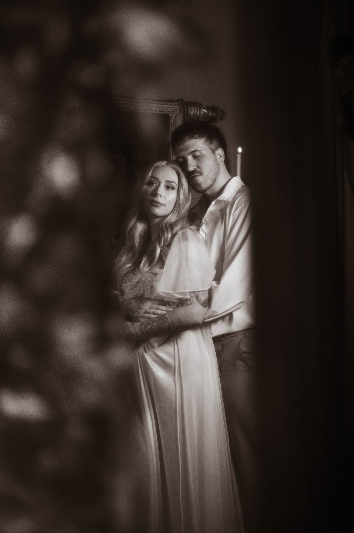 A romantic black and white portrait of a couple embracing in soft focus, surrounded by foliage and candlelight at Royal Lune Studio. The bride-to-be wears a flowing vintage-inspired gown, while her partner stands behind her in a ruffled shirt with his eyes closed. This artistic couple pose captures the moodiness and intimacy of dreamy engagement photography, ideal for Dallas engagement photos and romantic couples photography inspiration.