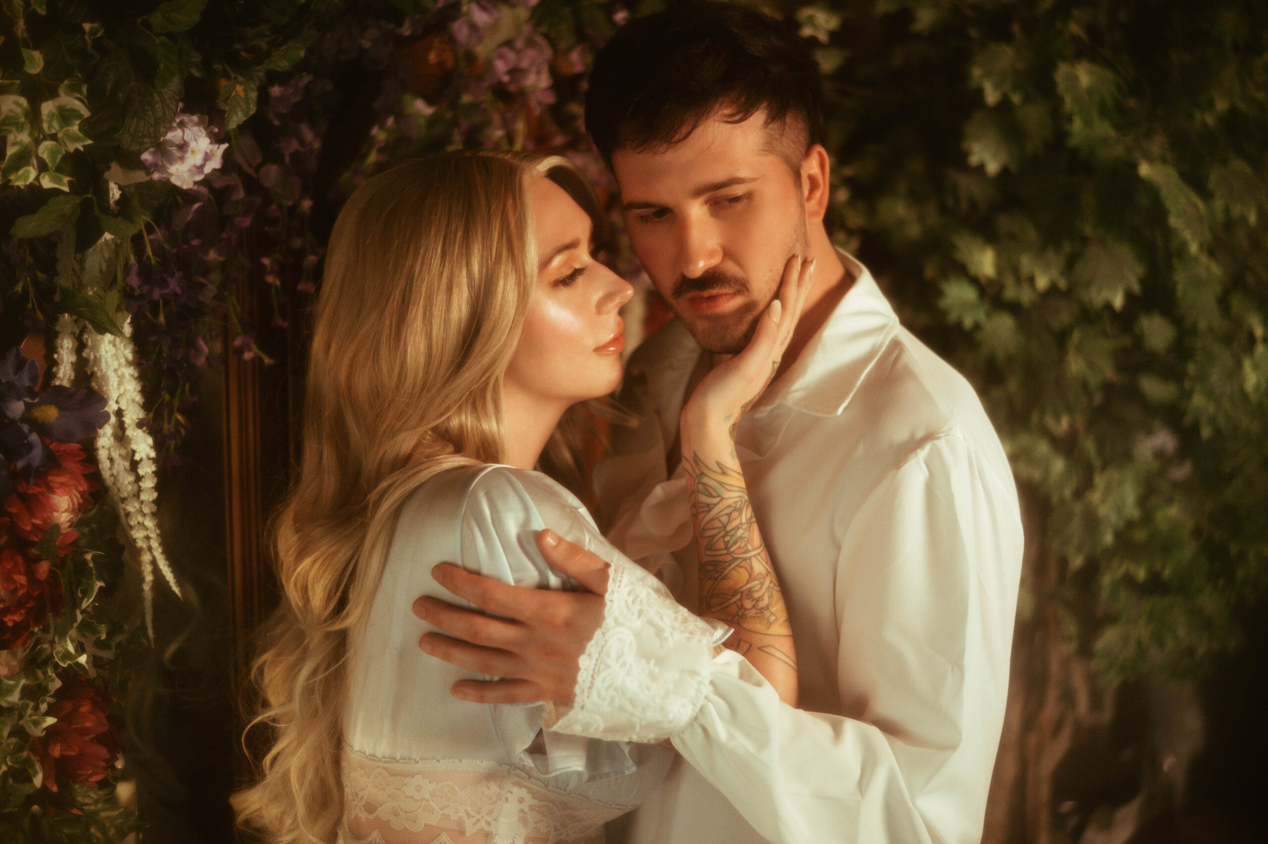 Romantic couple poses during a Dallas engagement photoshoot at Royal Lune Studio, with the woman in a vintage lace robe cupping her partner’s face under soft golden light. Surrounded by floral greenery, this dreamy engagement photography moment captures intimacy and tenderness, highlighting artistic couple poses and ethereal wardrobe styling.