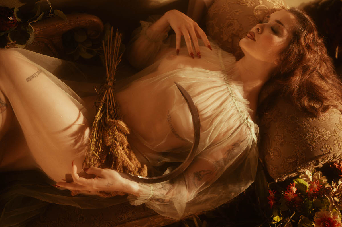 A fall boudoir photoshoot captures a woman reclining in sheer ivory fabric with a wheat bundle and harvest sycthe, surrounded by deep red florals in warm autumn light. Royal Lune Photo.