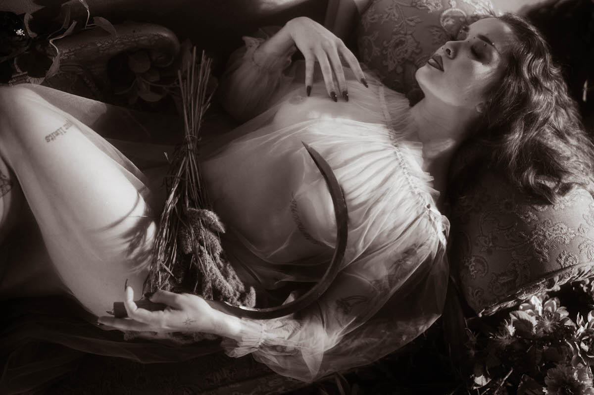 This moody fall photos black and white image features a woman reclining with a sickle and a bundle of dried wheat, recreating the pose of Allegory of Summer. Royal Lune Photo.