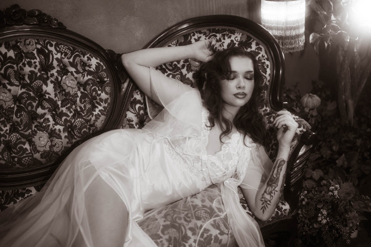 This moody fall photos black and white image shows a woman in sheer lace reclining on an antique tufted sofa with her arm raised above her head near a glowing fringe lamp. Royal Lune Photo.