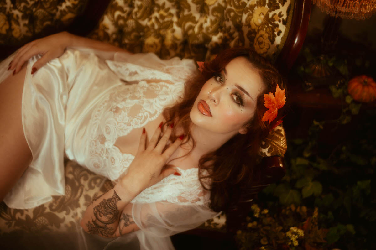 A fall boudoir photoshoot detail shot captures a woman in ivory satin and lace reclining with a maple leaf in her hair and floral tattoos visible on her arm. Royal Lune Photo.