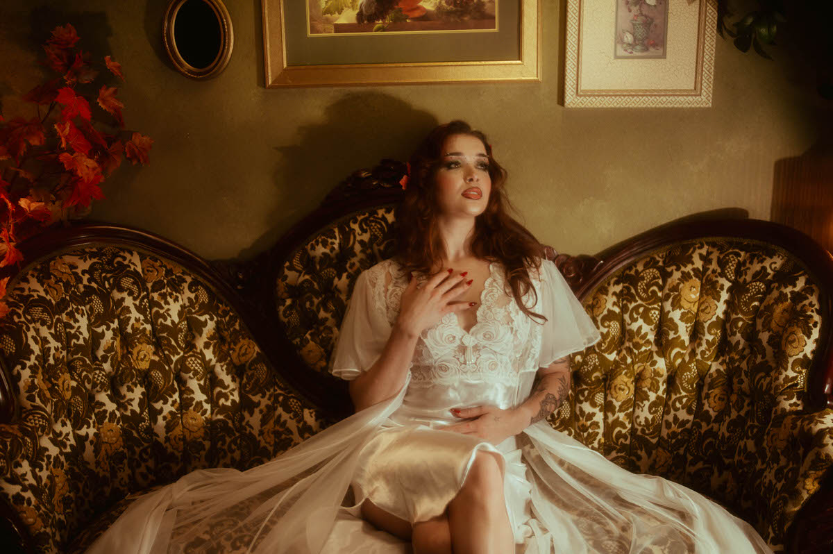 A fall photoshoot inspiration image shows a woman in a flowing ivory lace robe seated on an antique floral sofa beneath vintage gallery wall art. Royal Lune Photo.