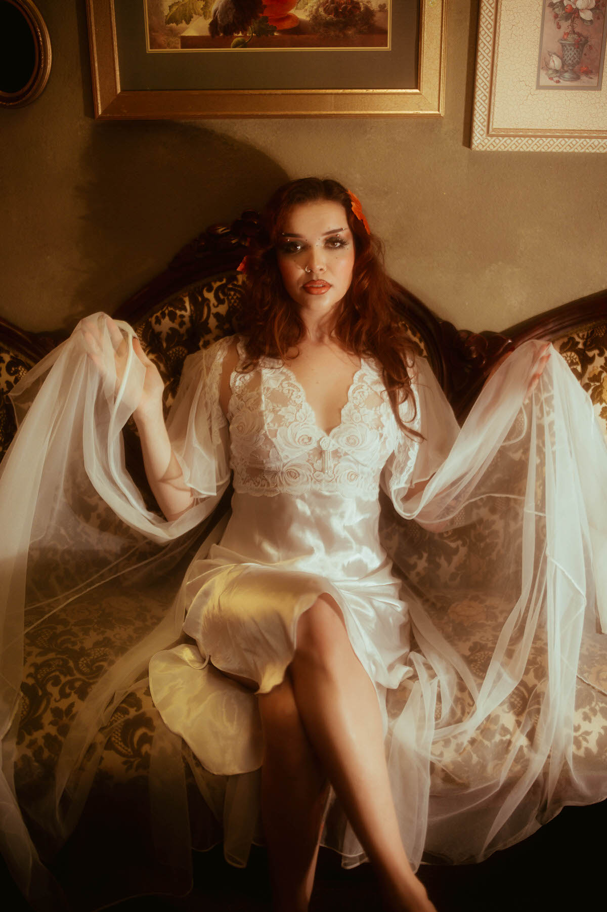 A fall boudoir photoshoot moment captures a woman in a satin and lace slip spreading sheer chiffon sleeves wide while seated on a vintage damask sofa. Royal Lune Photo.