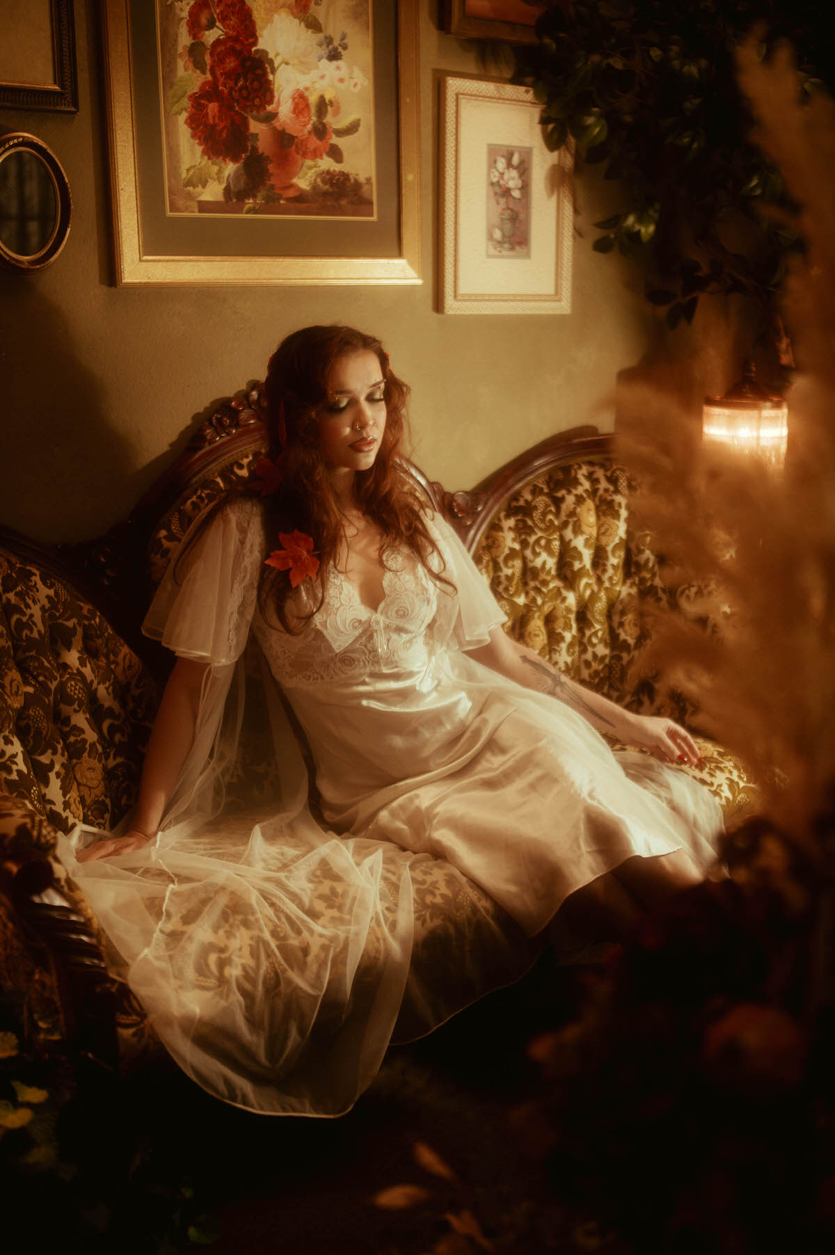 A fall photoshoot inspiration scene shows a woman in a flowing satin gown with a maple leaf tucked into her curls, lit by warm lantern glow on an antique sofa. Royal Lune Photo.