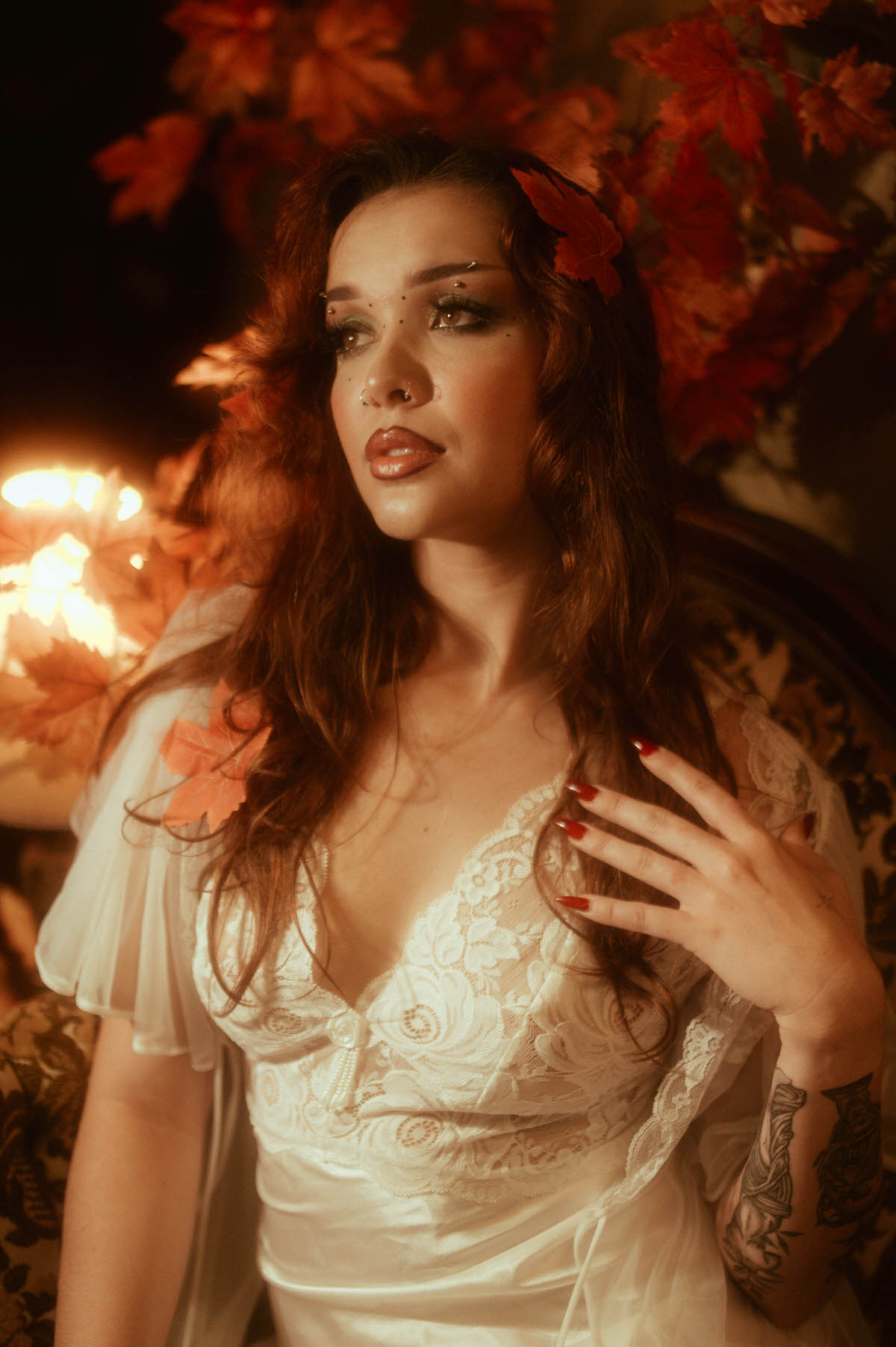 This moody fall photos close up portrait highlights a woman in white lace with autumn leaves woven through her hair, candlelight flickering beside her. Royal Lune Photo.