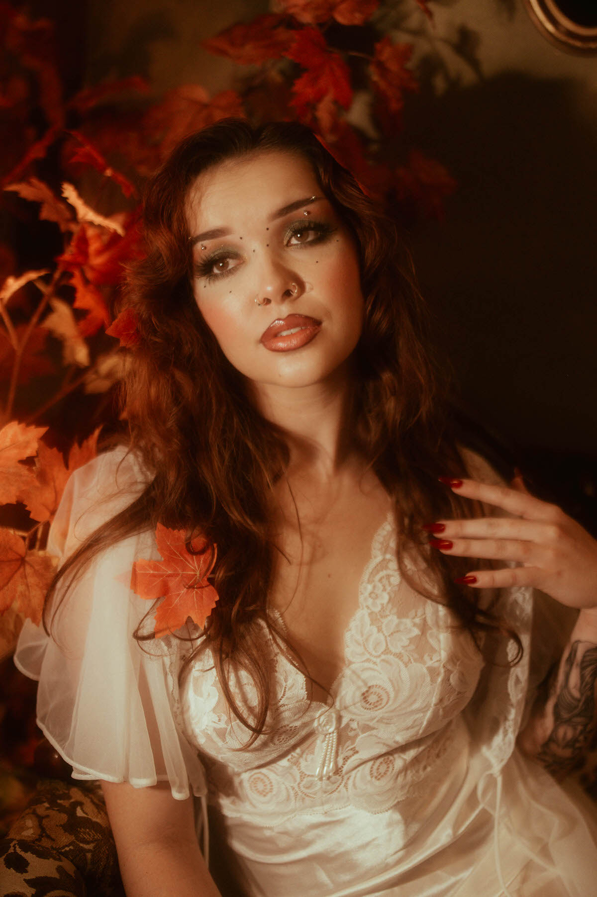 Dreamy autumn photography highlights a woman in white lace lingerie nestled among vibrant red maple leaves, her gaze soft and her curls catching the golden light. Royal Lune Photo.
