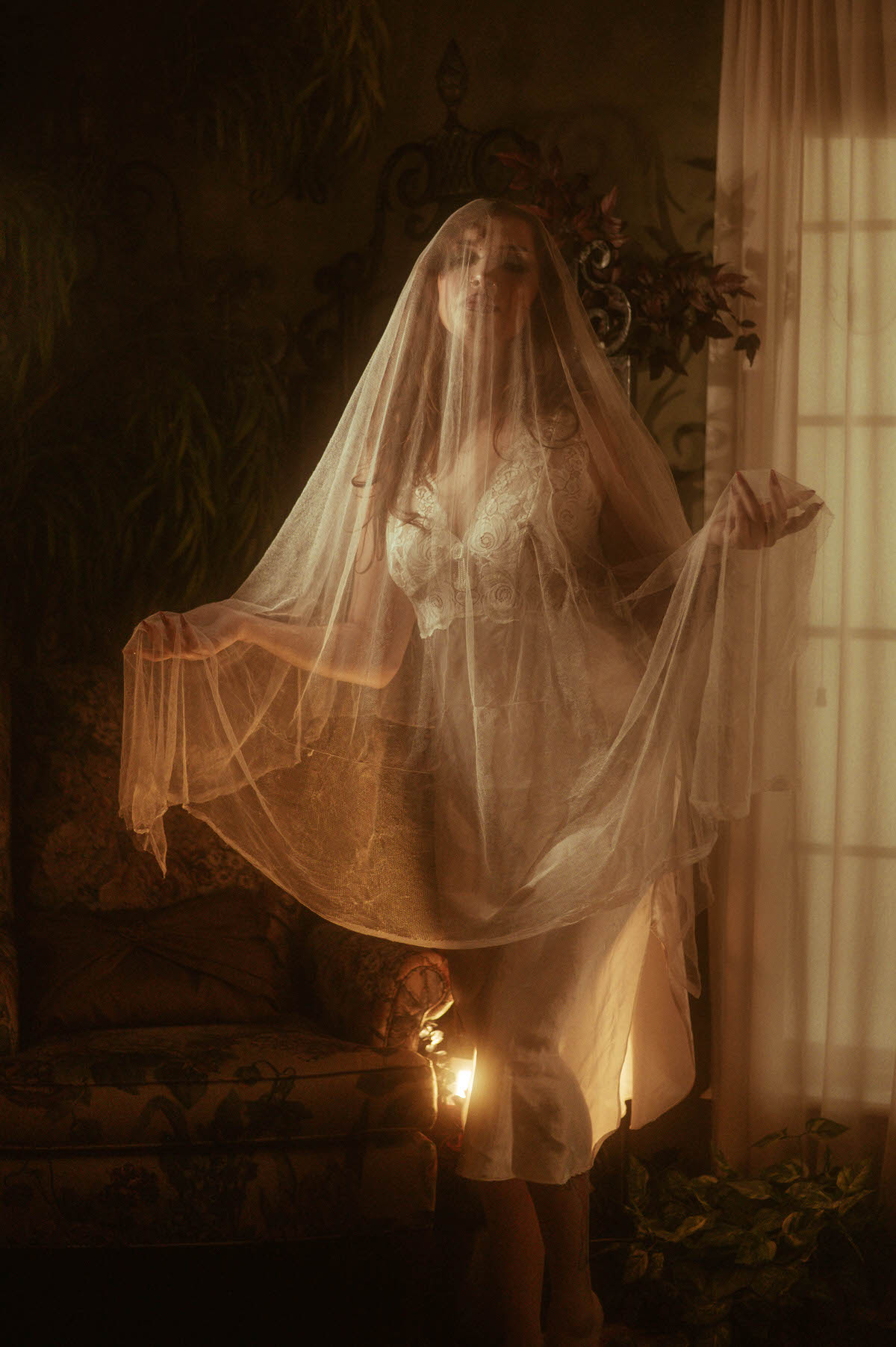 This dreamy autumn photography image shows a woman veiled in flowing tulle standing beside a candlelit window with sheer curtains. Royal Lune Photo.