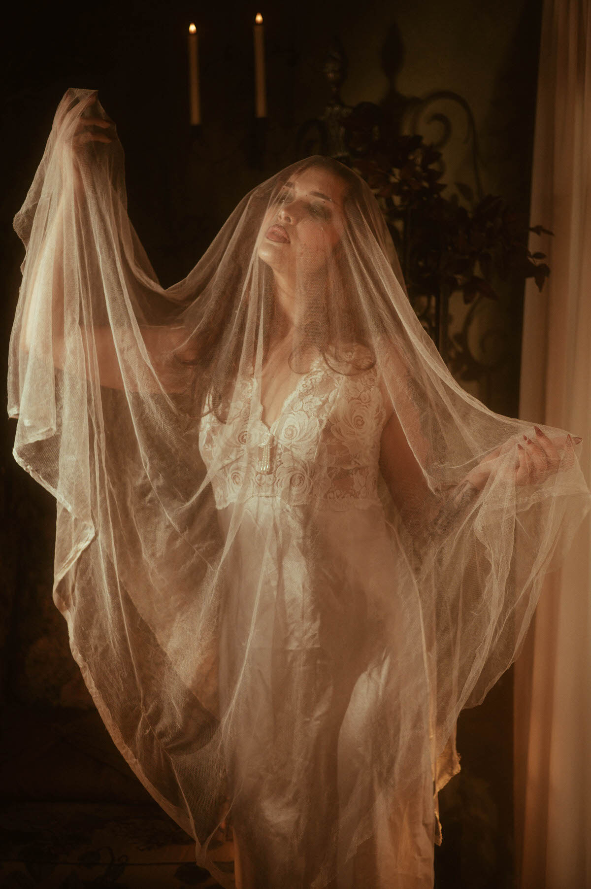 Fall photoshoot inspiration highlights a woman in white lace lifting a sheer veil overhead as candlelight flickers in the background. Royal Lune Photo.