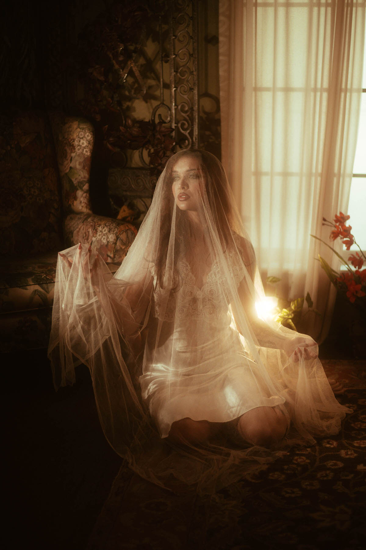 A fall boudoir photoshoot scene shows a woman veiled in tulle seated by a sunlit window with red florals and soft golden light around her. Royal Lune Photo.