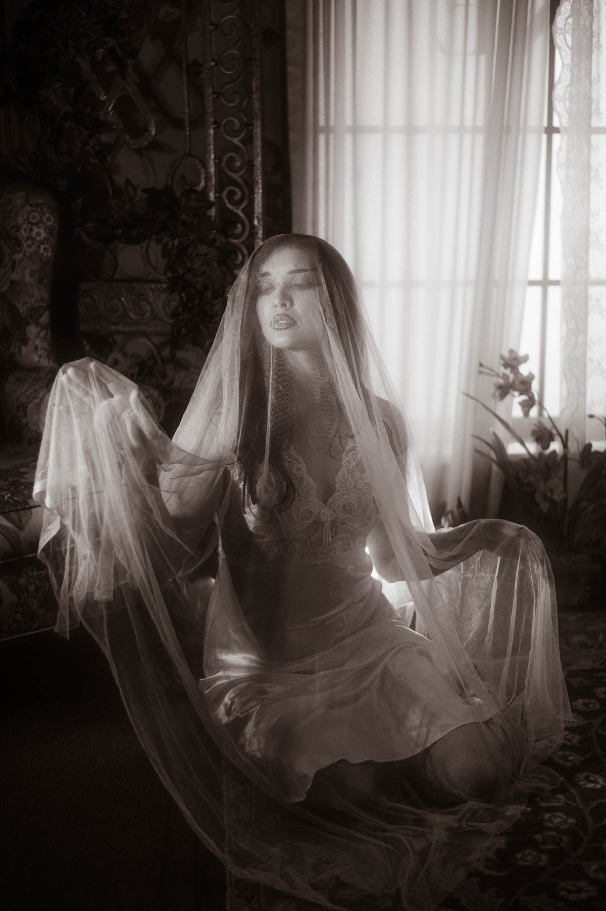 This moody fall photos black and white image features a woman kneeling in a veil and lace gown beside a sunlit window with sheer curtains. Royal Lune Photo.
