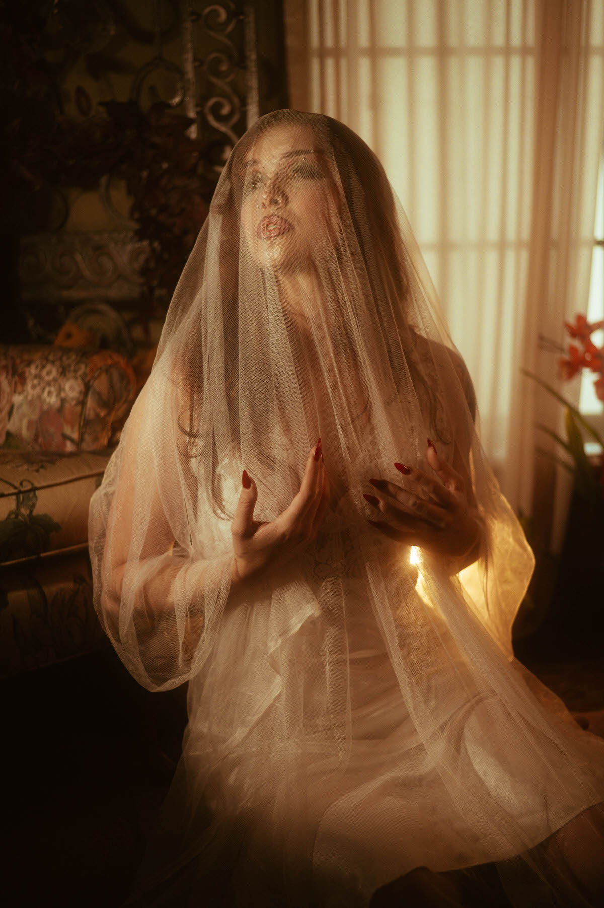 A fall boudoir photoshoot moment captures a woman cloaked in sheer tulle with her hands pressed gently to her chest beside a sunlit window. Royal Lune Photo.