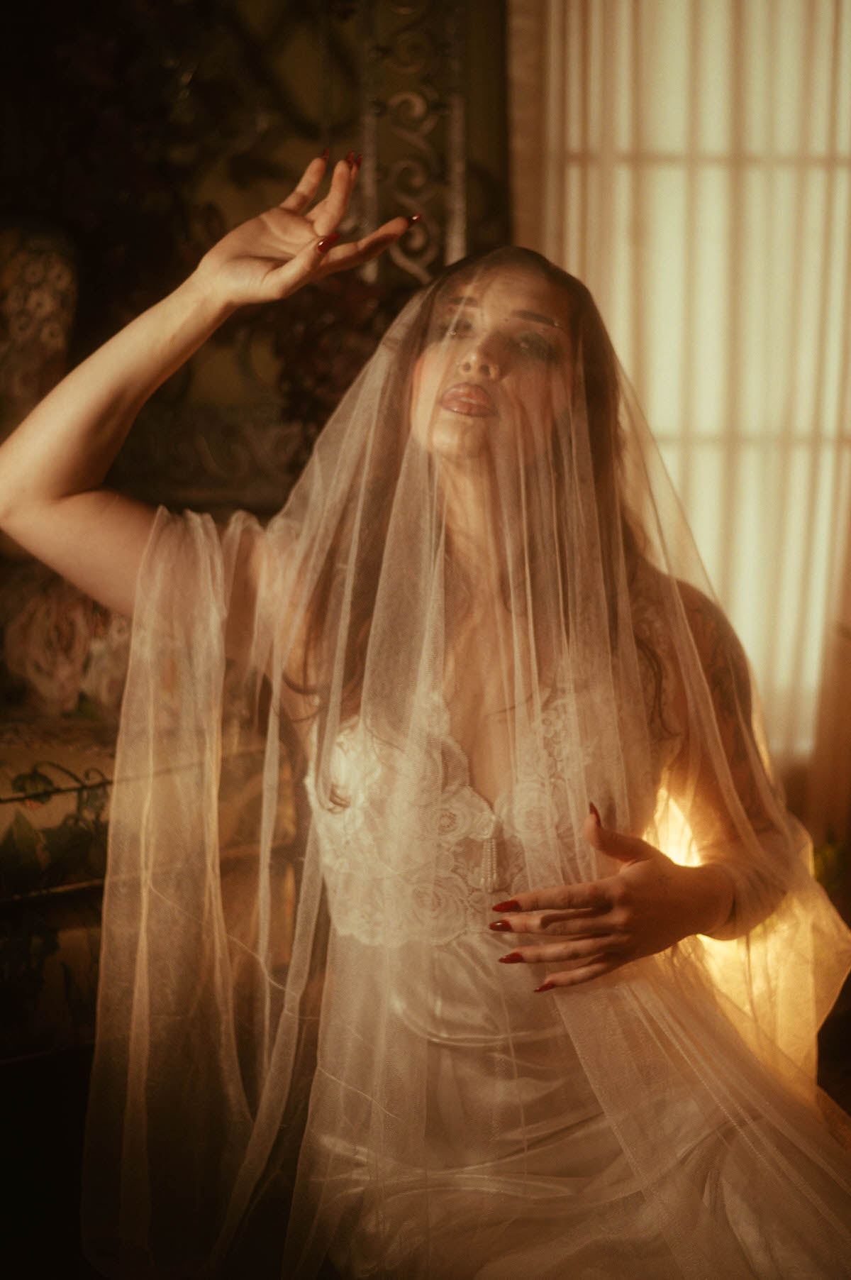 Dreamy autumn photography shows a woman veiled in flowing tulle with one hand raised gracefully against warm golden window light. Royal Lune Photo.