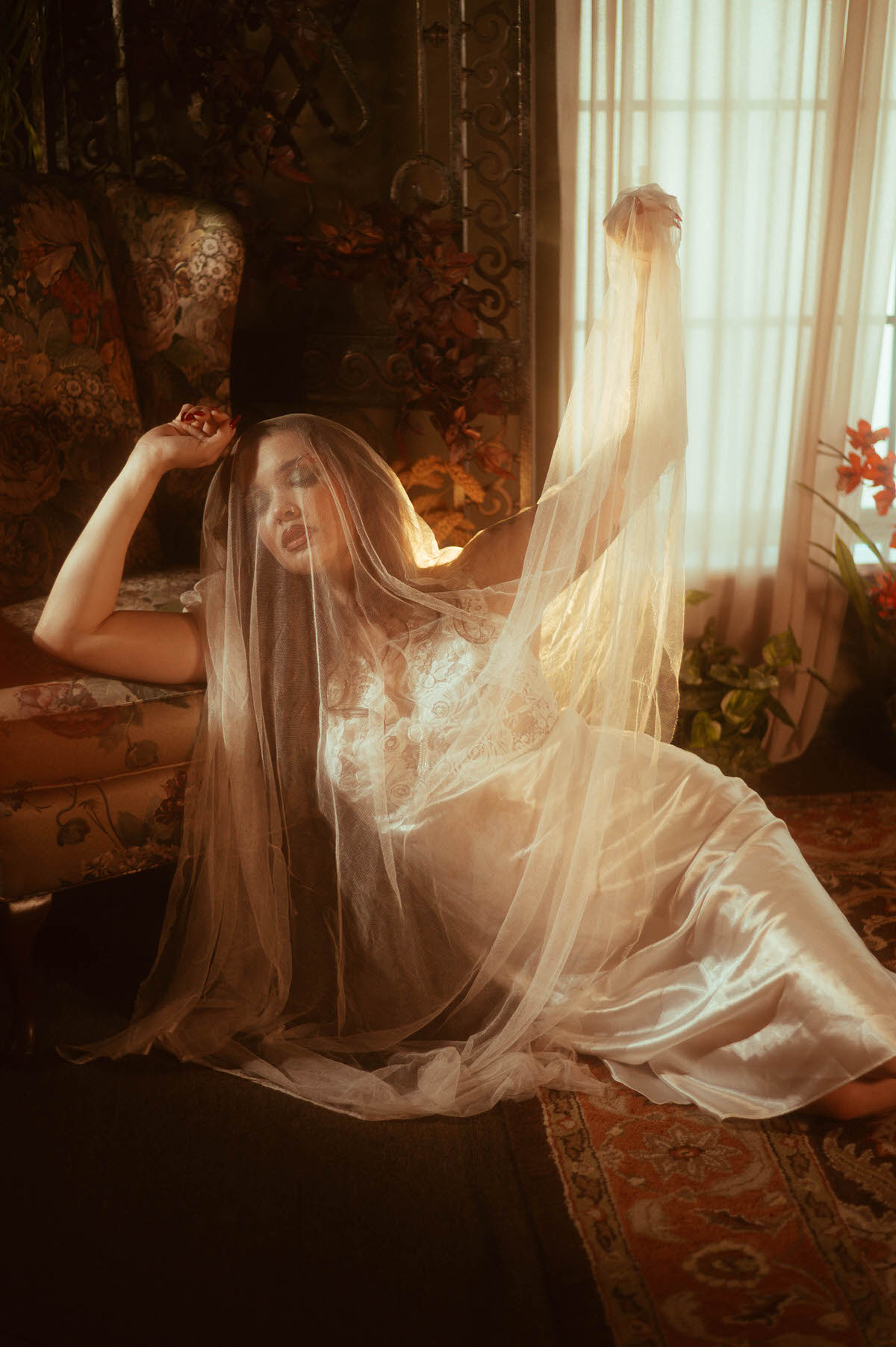 A fall photoshoot inspiration scene features a woman reclining on a floral antique sofa with a sheer veil lifted toward sunlit curtains. Royal Lune Photo.