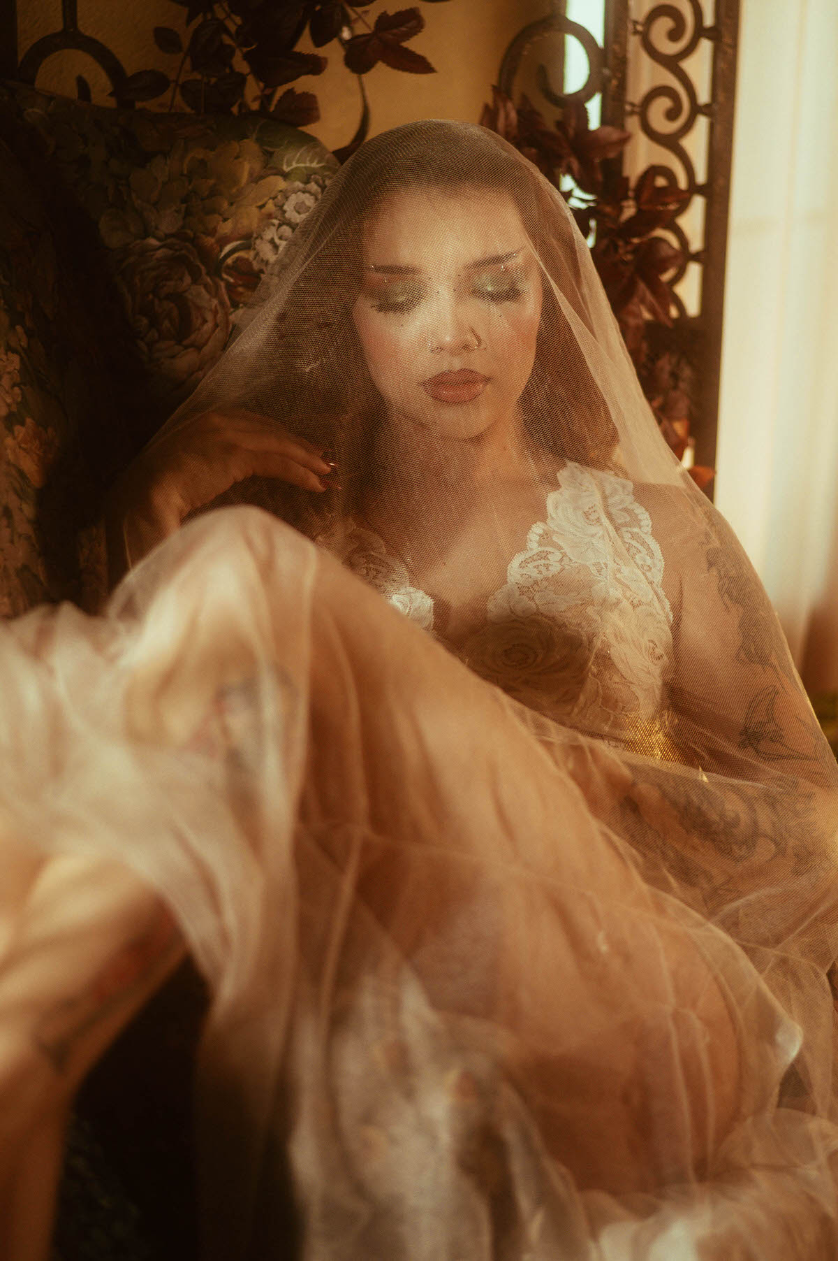 A fall boudoir photoshoot close up captures a woman in white lace veiled in tulle with soft glittering makeup and warm sunlight filtering through wrought iron decor. Royal Lune Photo.