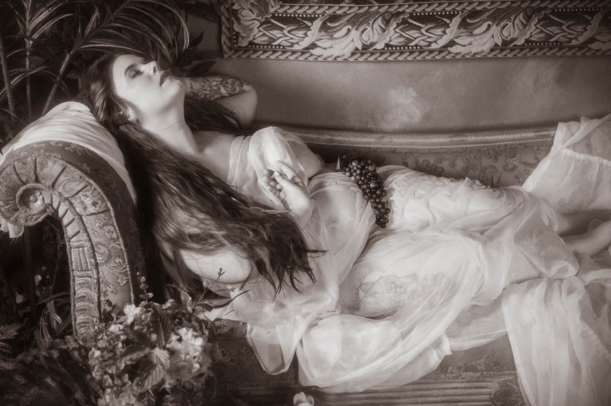 Black and white image of a reclining woman in sheer drapery on a vintage couch, holding grapes with eyes closed, evoking Pre-Raphaelite boudoir and classical still-life beauty.