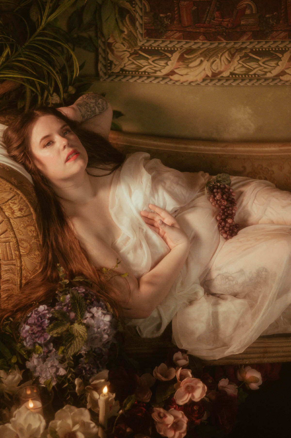 Reclining plus-sized woman draped in sheer white fabric with grapes on her hip, surrounded by candles and lush florals, channeling a romantic Pre-Raphaelite boudoir mood.