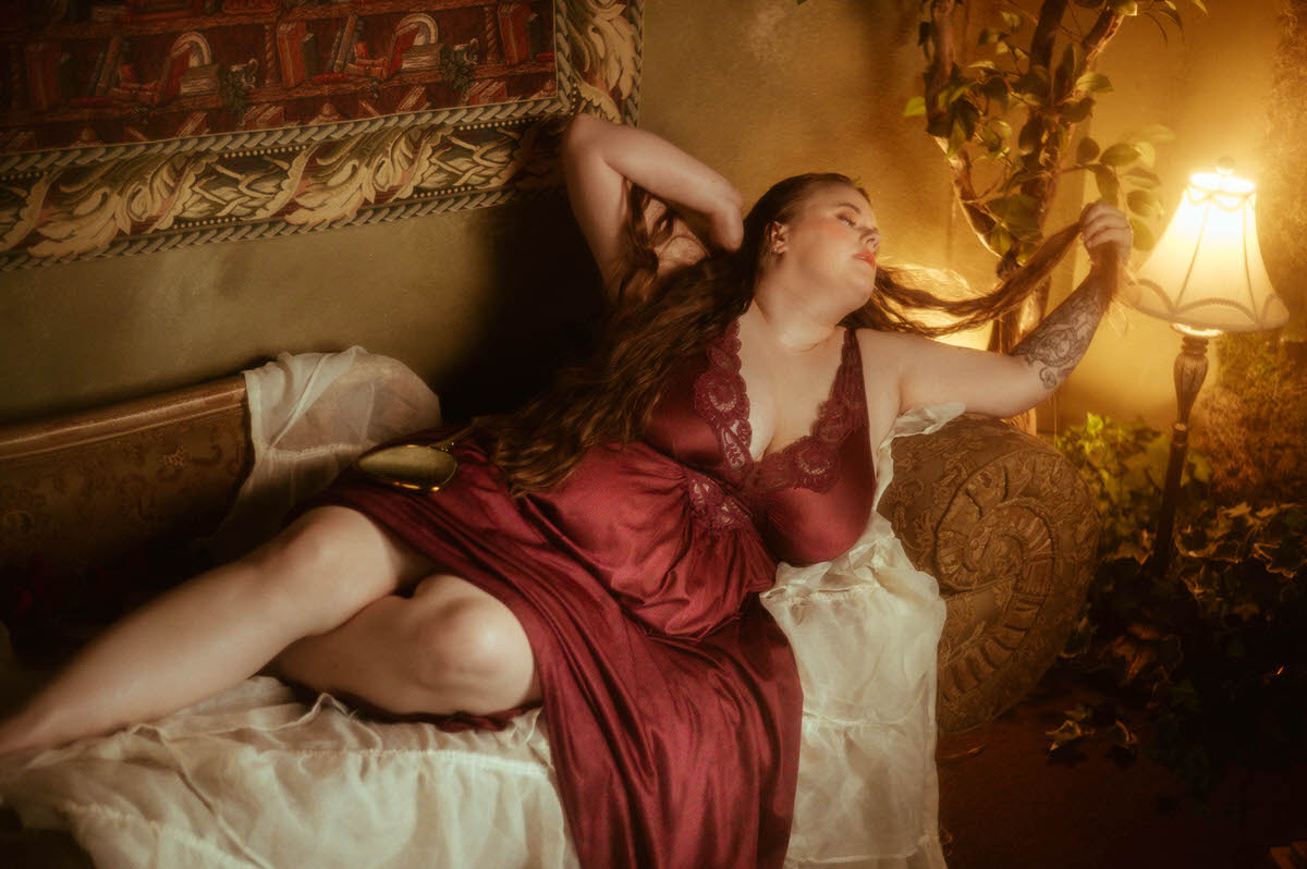 A woman in a burgundy vintage Olga nightgown lounges on an antique couch, lit by warm lamplight, running her fingers through her hair in a lush, pre-Raphaelite boudoir scene at a Dallas boudoir studio.