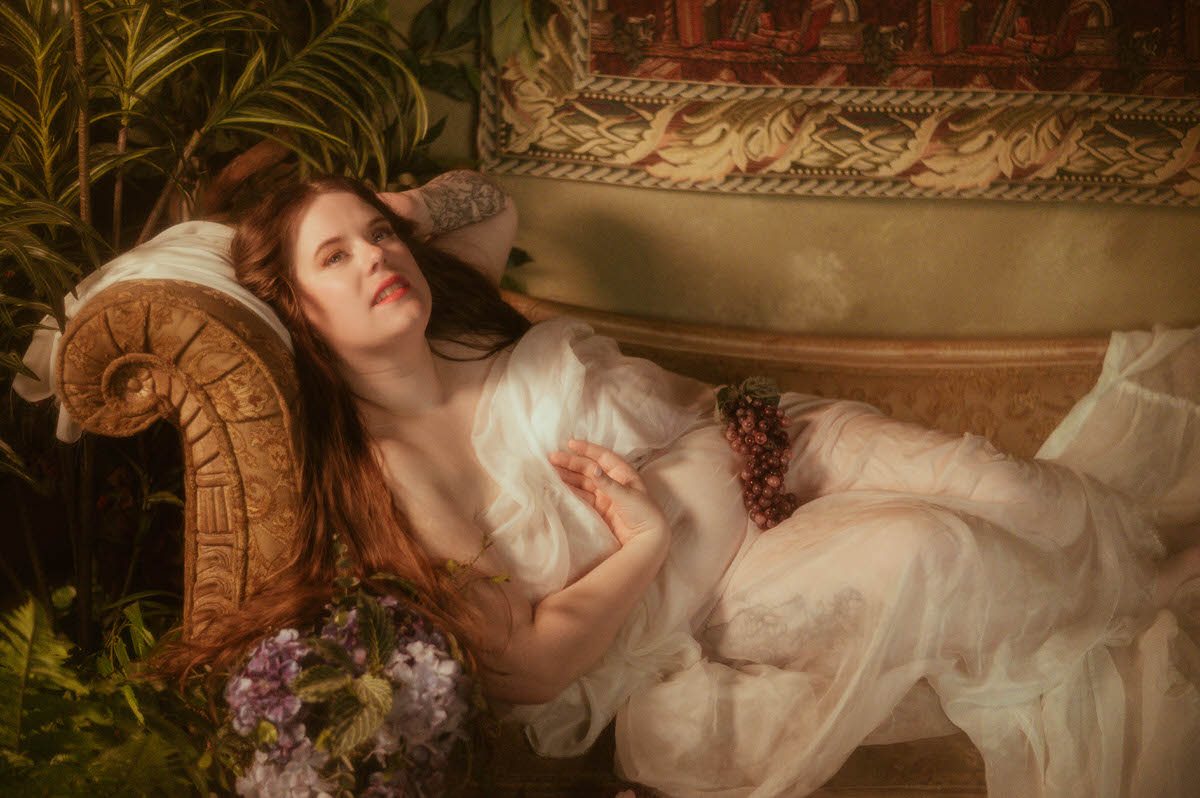 Romantic Pre-Raphaelite boudoir scene of a reclining plus-sized woman in sheer drapery, resting on a vintage couch with grapes on her hip, surrounded by greenery and florals.