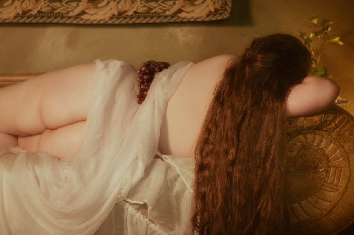 Back view of a plus-sized woman lying on a vintage chaise draped in sheer white fabric, with long auburn hair cascading down and grapes resting on her hip in a dreamy Pre-Raphaelite boudoir scene.
