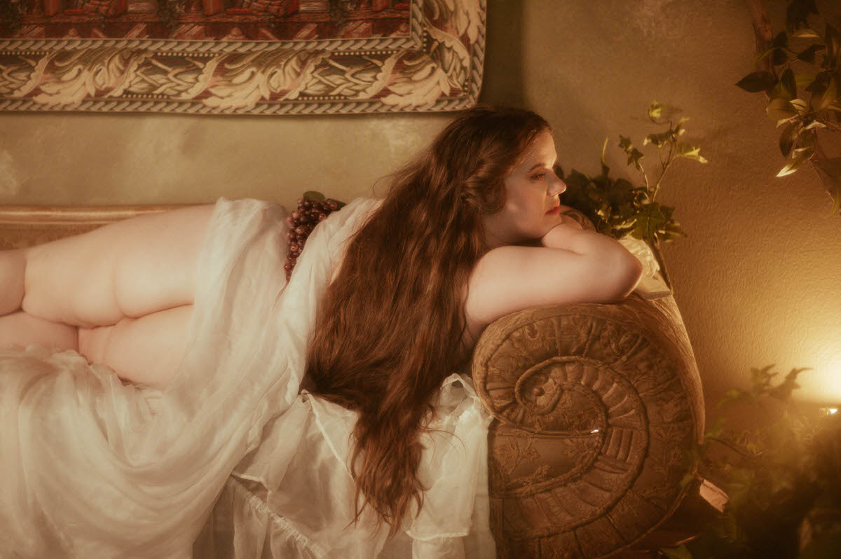 A curvy plus-sized woman reclines on a scroll-armed vintage sofa draped in sheer white fabric, her long wavy hair flowing down her back as she gazes softly into warm candlelight, with grapes resting on her hip in a romantic Pre-Raphaelite boudoir setting.