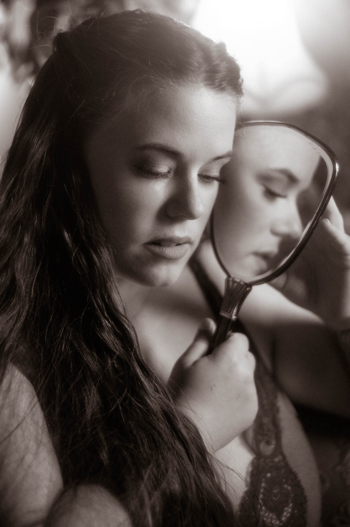 Black and white portrait of a woman in a lace vintage nightgown holding a hand mirror, her downcast eyes and reflection evoking soft emotion in a pre-Raphaelite boudoir session at a Dallas boudoir studio.
