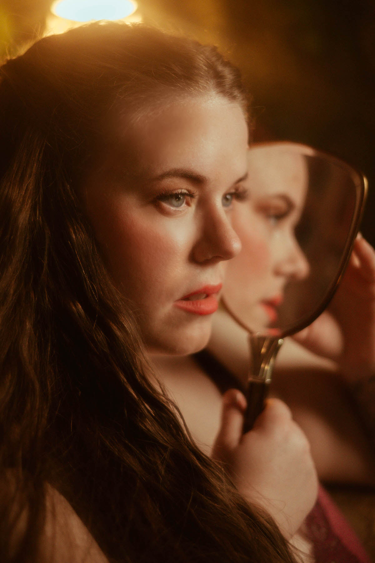 A close-up pre-Raphaelite boudoir portrait of a woman gazing into an antique hand mirror, her reflection softly glowing under golden light at a Dallas boudoir studio.