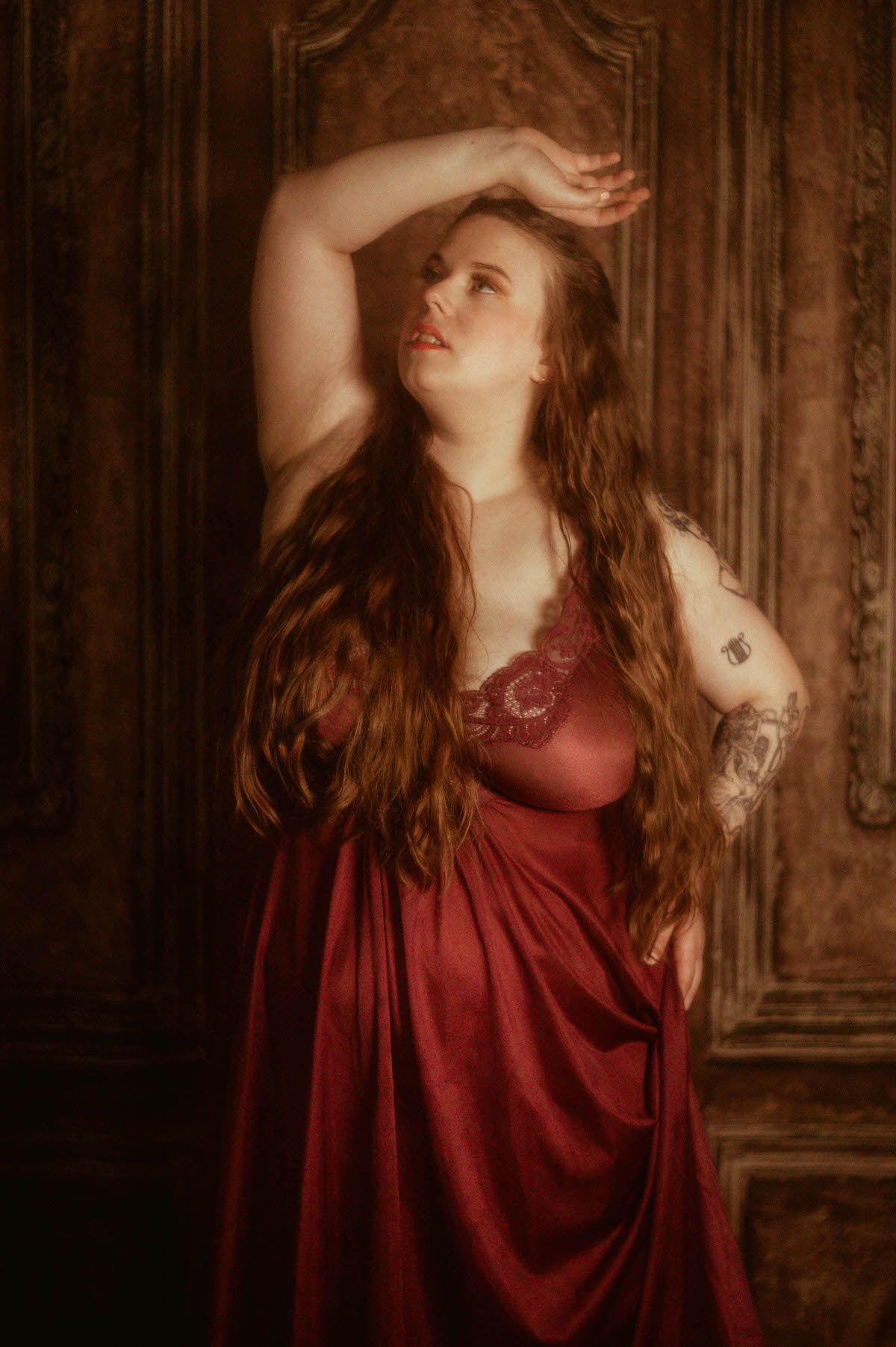 A woman in a flowing burgundy vintage Olga nightgown stands gracefully with her arm arched overhead, channeling pre-Raphaelite beauty in a fine art boudoir session at a Dallas boudoir studio.
