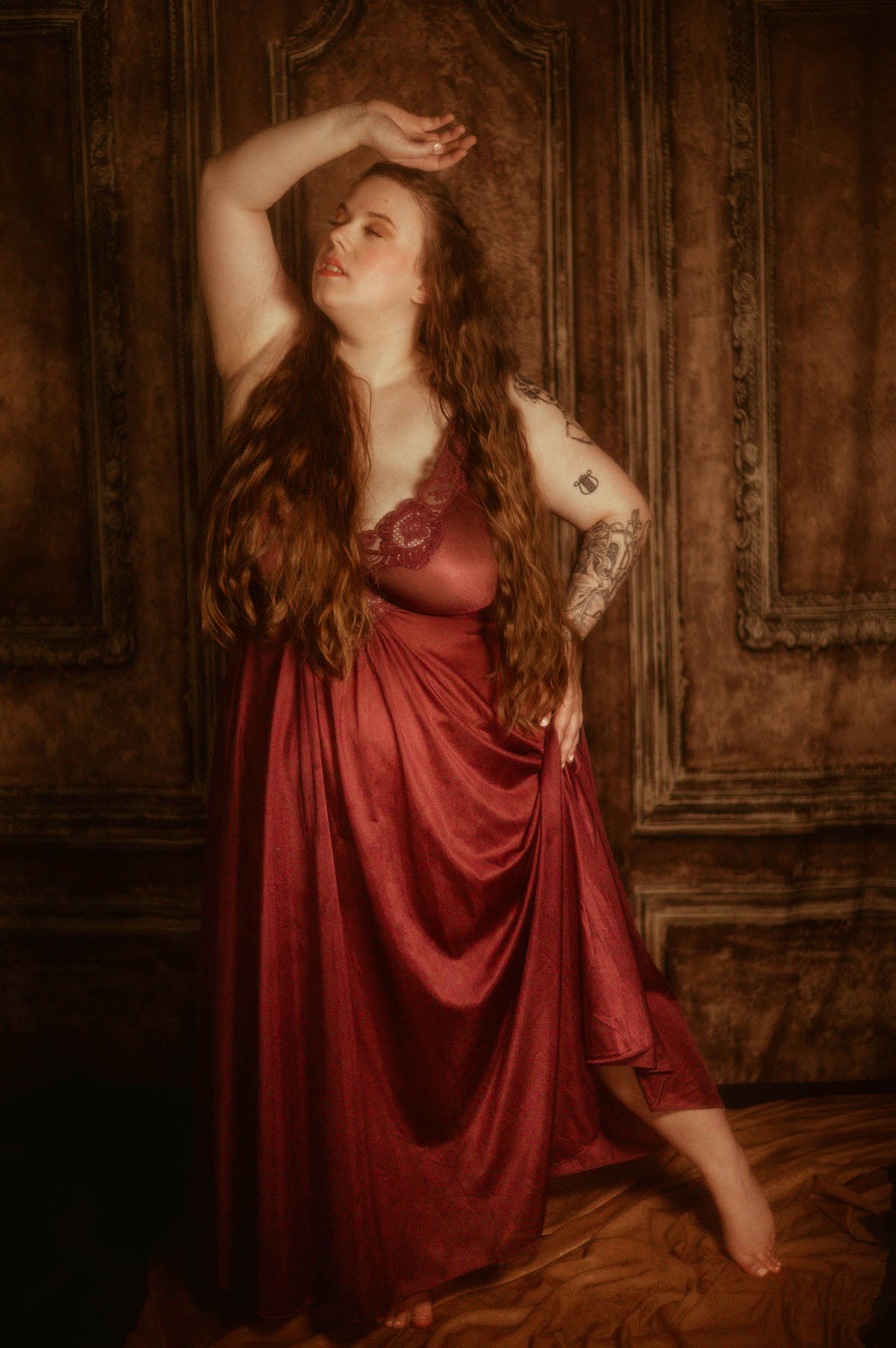 A woman in a flowing burgundy vintage Olga nightgown poses barefoot with one arm raised in front of an ornate backdrop, embodying fine art boudoir and pre-Raphaelite elegance at a Dallas boudoir studio.