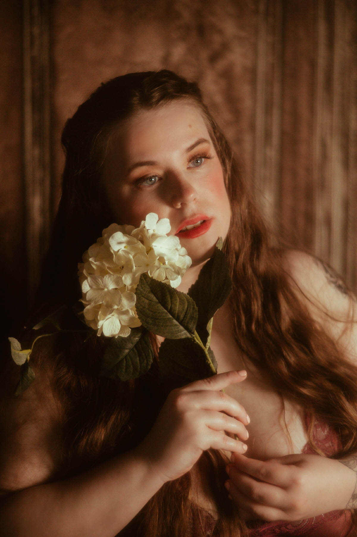 Close-up fine art boudoir portrait of a woman with long, wavy hair holding a white hydrangea near her face, styled in a soft pre-Raphaelite aesthetic with dreamy lighting at a Dallas boudoir studio.