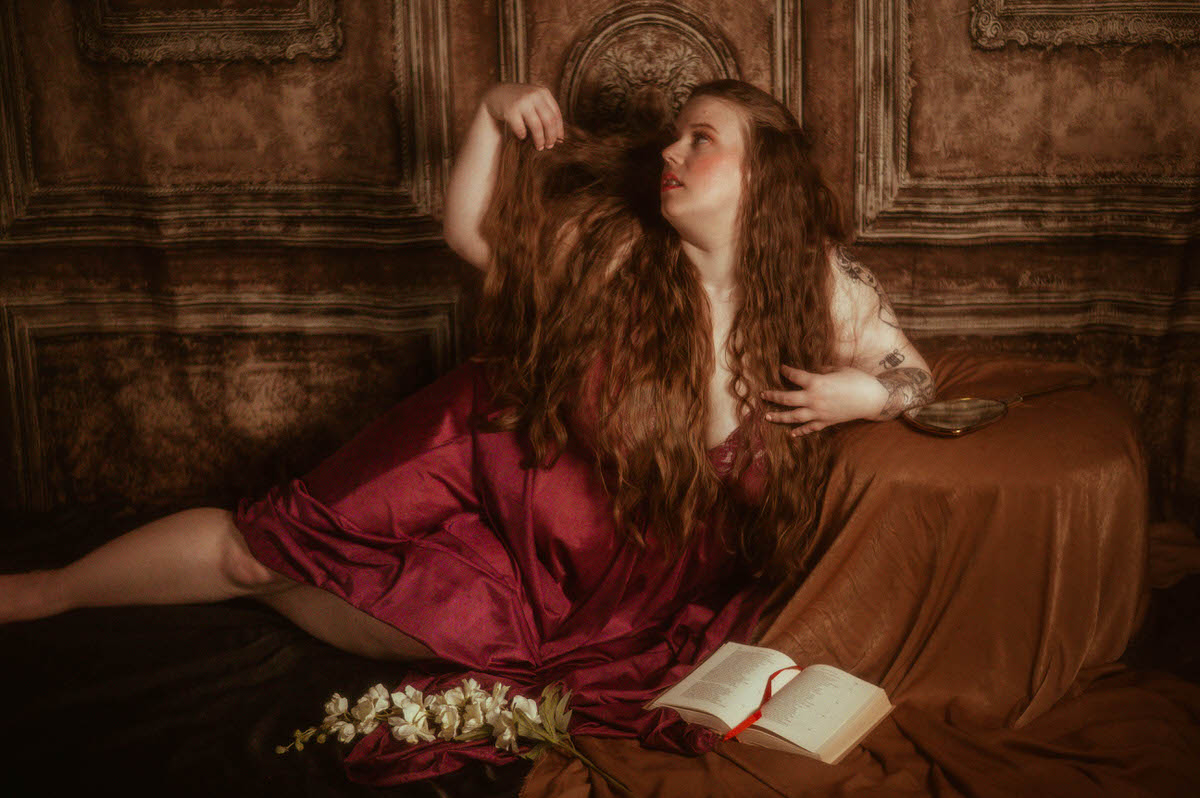 A woman reclines on draped fabric in a burgundy vintage Olga nightgown, holding her long hair while surrounded by white flowers, an open book, and a mirror in a poetic pre-Raphaelite boudoir scene at a Dallas boudoir studio.
