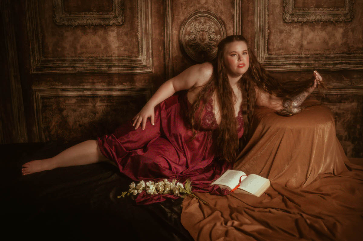 A woman in a burgundy nylon Olga nightgown reclines against a brown velvet backdrop, holding her long hair beside a vintage mirror, open book, and white florals in a fine art pre-Raphaelite boudoir session at a Dallas boudoir studio.