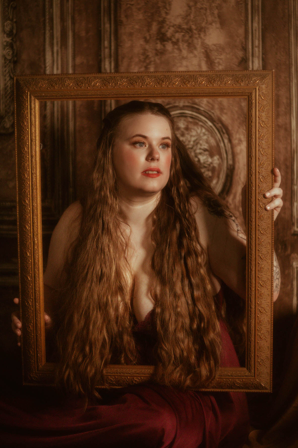 A woman with long wavy hair gazes thoughtfully while holding an ornate picture frame in front of her chest, styled in a vintage nightgown for a fine art pre-Raphaelite boudoir session at a Dallas boudoir studio.