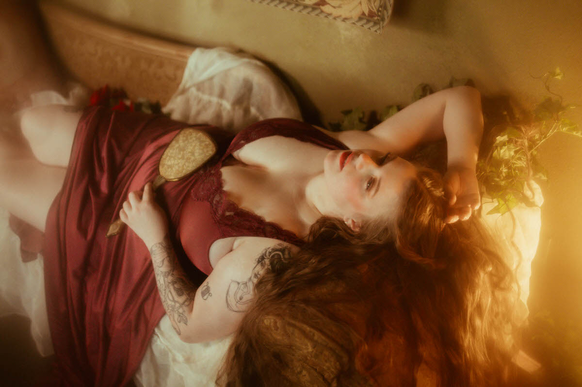 A woman reclines on a vintage couch in a burgundy lace nightgown, holding an antique hand mirror with her long hair fanned out, evoking a dreamy pre-Raphaelite boudoir aesthetic at a Dallas boudoir studio.