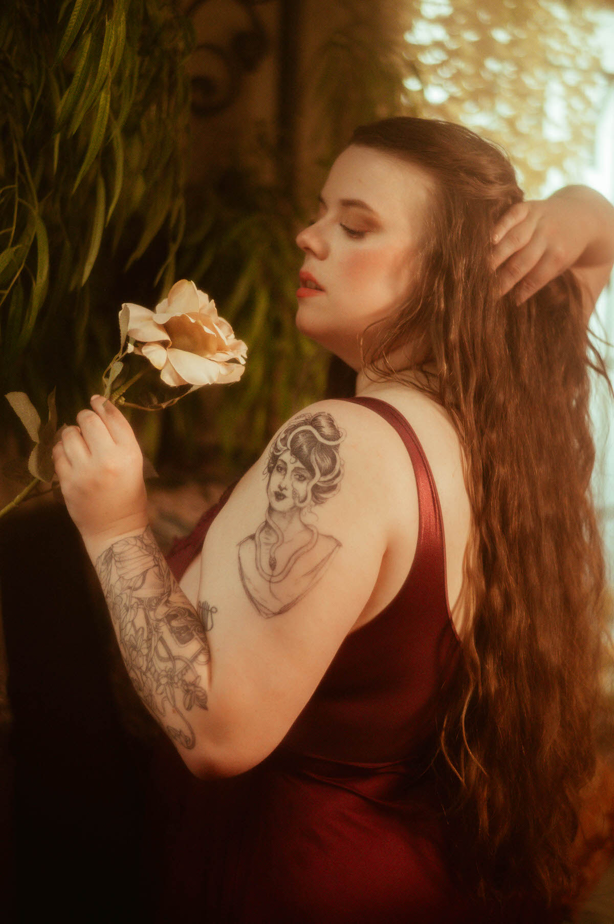 A tattooed woman in a burgundy vintage nightgown holds a peach-toned rose and touches her hair, surrounded by soft greenery in a romantic pre-Raphaelite boudoir scene at a Dallas boudoir studio.