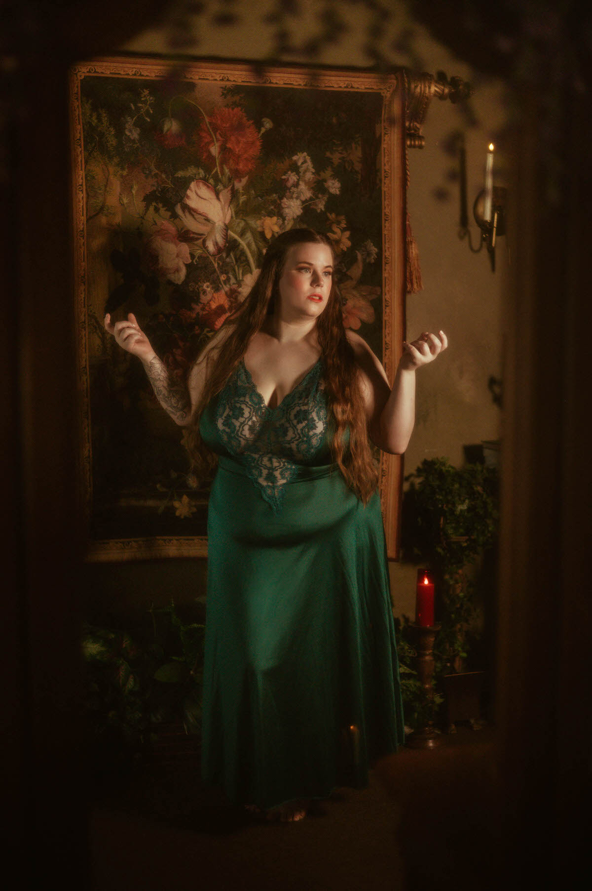 A woman in a flowing vintage teal lace gown stands barefoot in front of a floral tapestry, bathed in candlelight, evoking ethereal pre-Raphaelite boudoir styling at a Dallas boudoir studio.