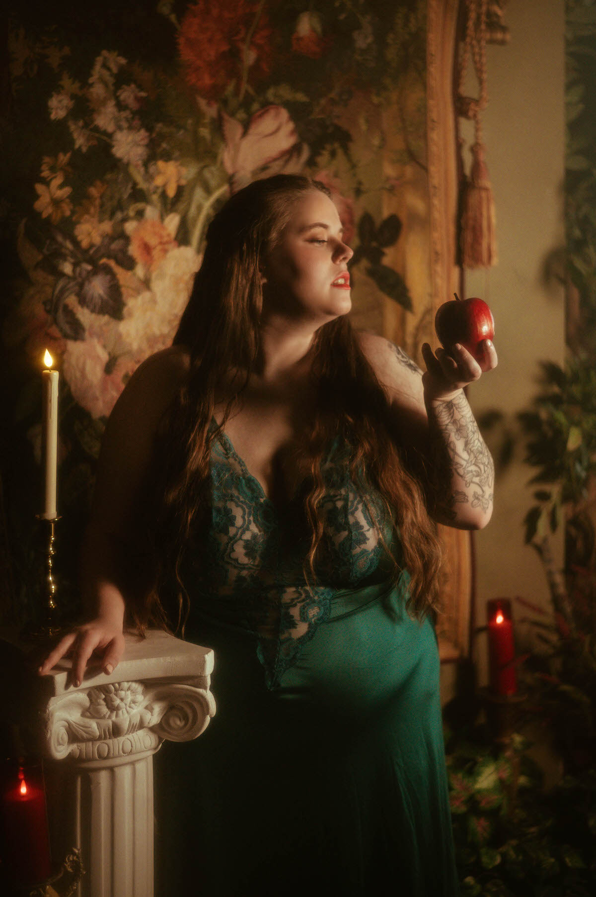 A woman in a teal lace vintage gown holds a red apple while standing beside a white column and lit candles, styled in a dark floral pre-Raphaelite boudoir scene at a Dallas boudoir studio.