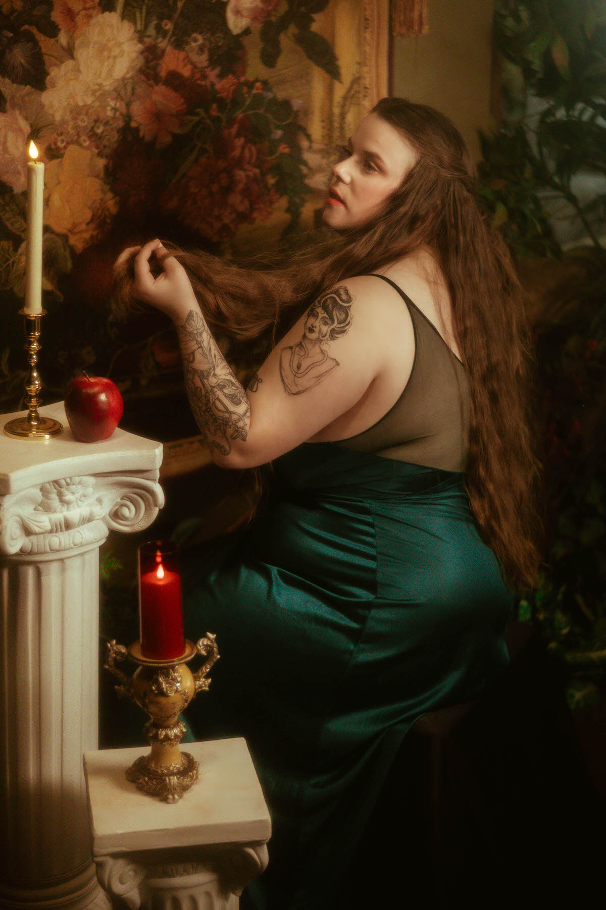 A woman in a vintage teal nightgown sits beside lit candles and a red apple, running her fingers through her hair in front of a floral tapestry, styled in a moody pre-Raphaelite boudoir session at a Dallas boudoir studio.