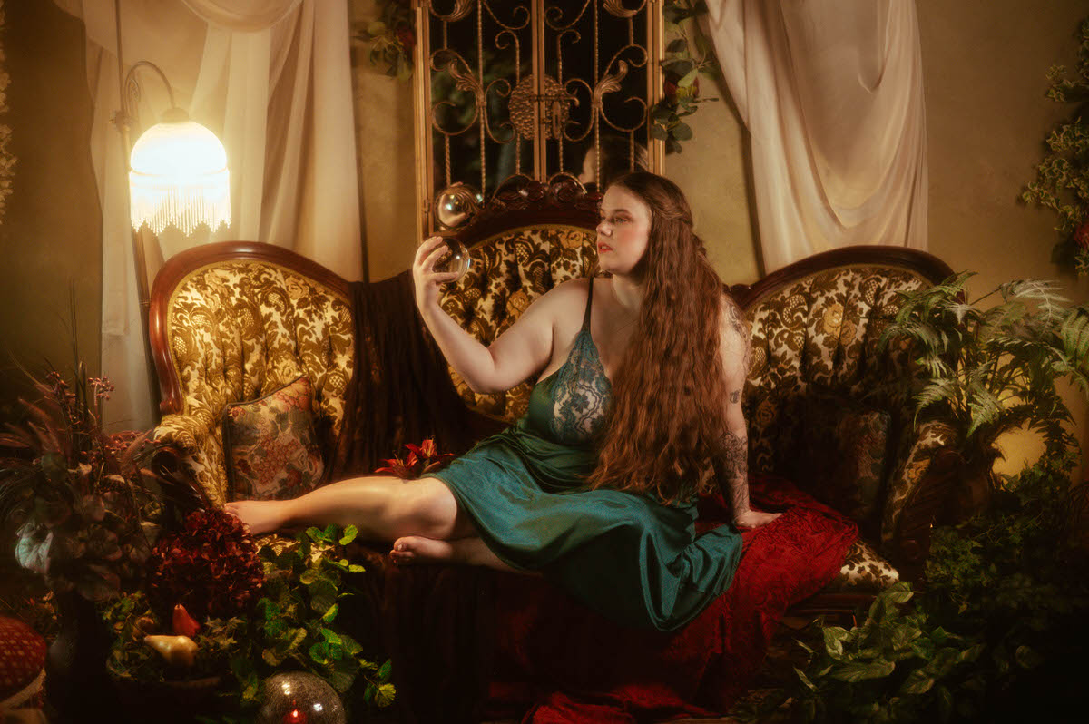A woman in a vintage teal nightgown lounges on a vintage settee, holding a glass orb surrounded by lush greenery, velvet textures, and antique decor in a pre-Raphaelite boudoir session at a Dallas boudoir studio.
