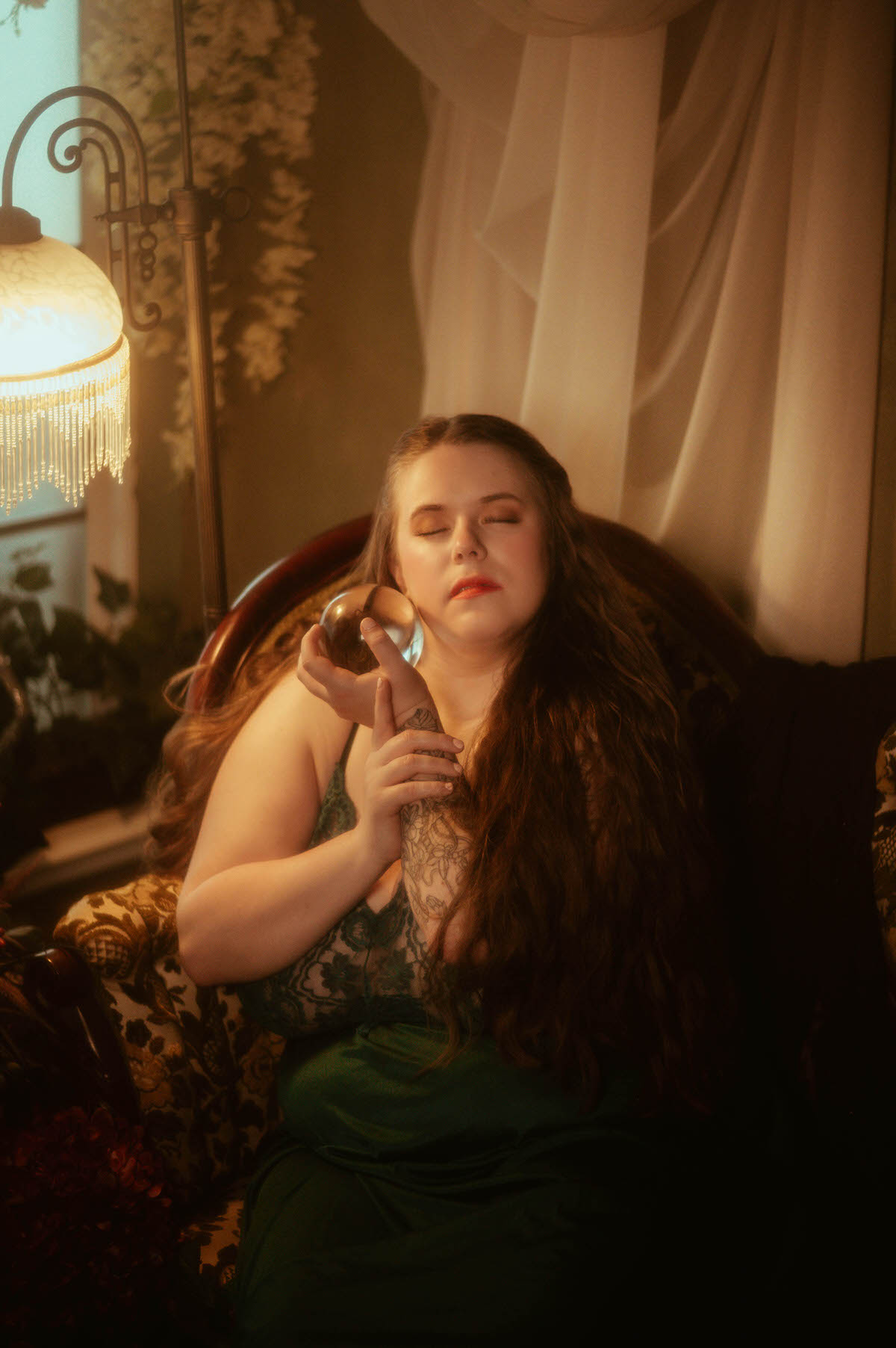 A woman in a vintage teal nightgown sits with eyes closed, gently holding a glass orb near her cheek under vintage lamplight, styled in a dreamy pre-Raphaelite boudoir session at a Dallas boudoir studio.