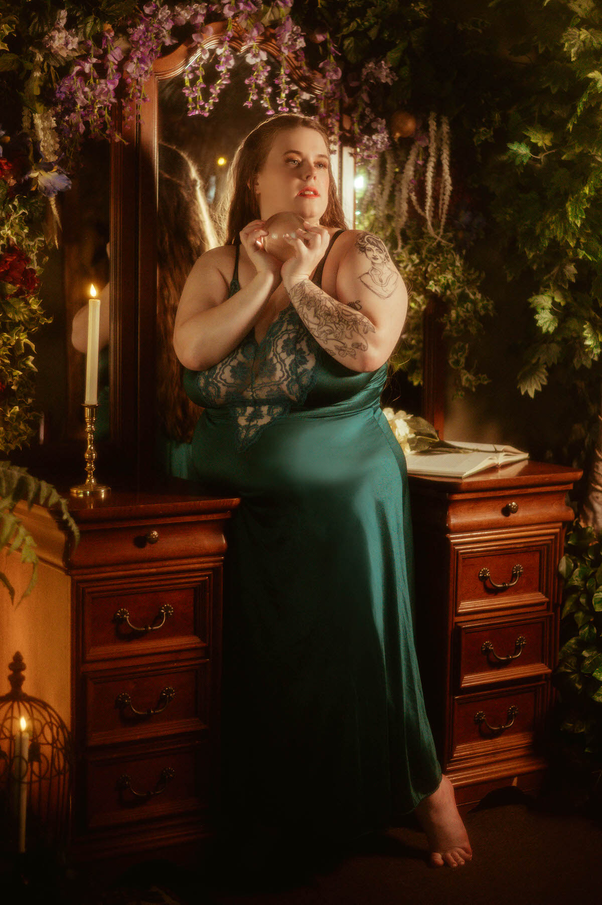 A curvy woman in a vintage teal nightgown sits at a vintage vanity surrounded by ivy, florals, and candlelight, holding a glass orb during a pre-Raphaelite boudoir session at a Dallas boudoir studio.