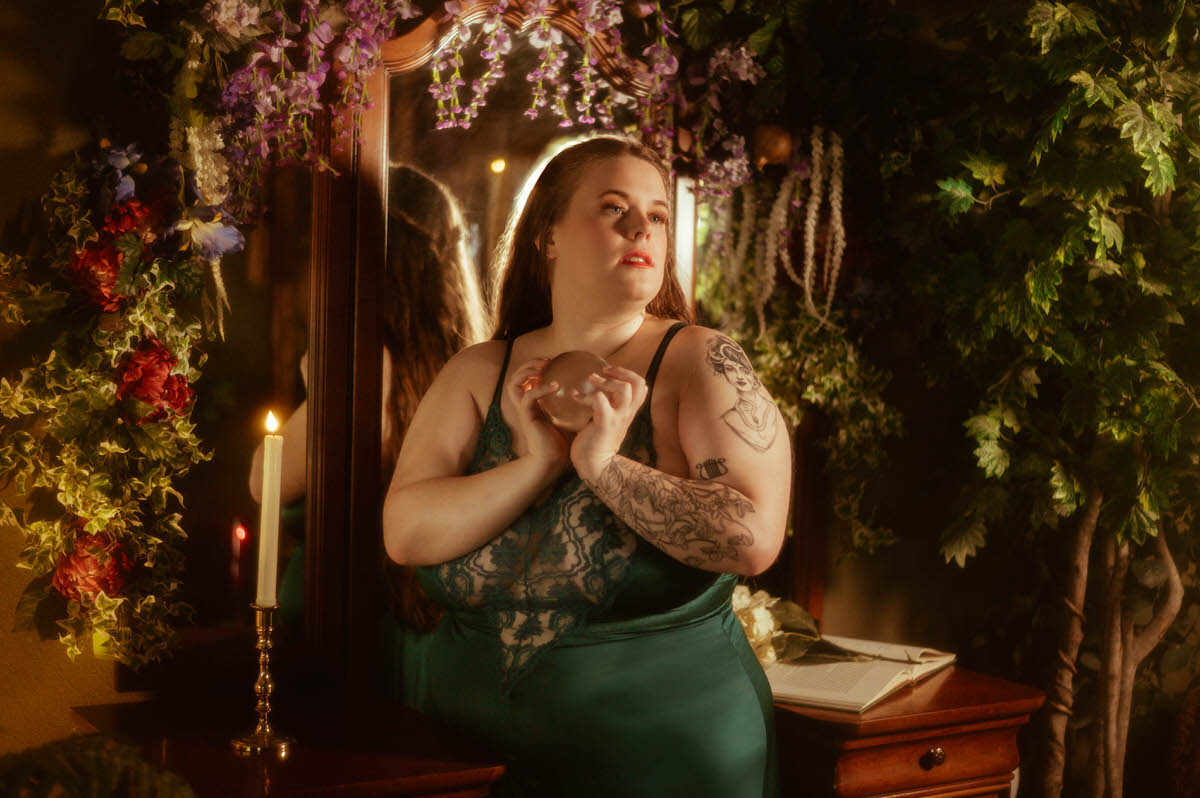 A woman in a teal lace gown stands before a mirror framed in florals, holding a glass orb close to her chest in a romantic pre-Raphaelite boudoir setting at a Dallas boudoir studio.
